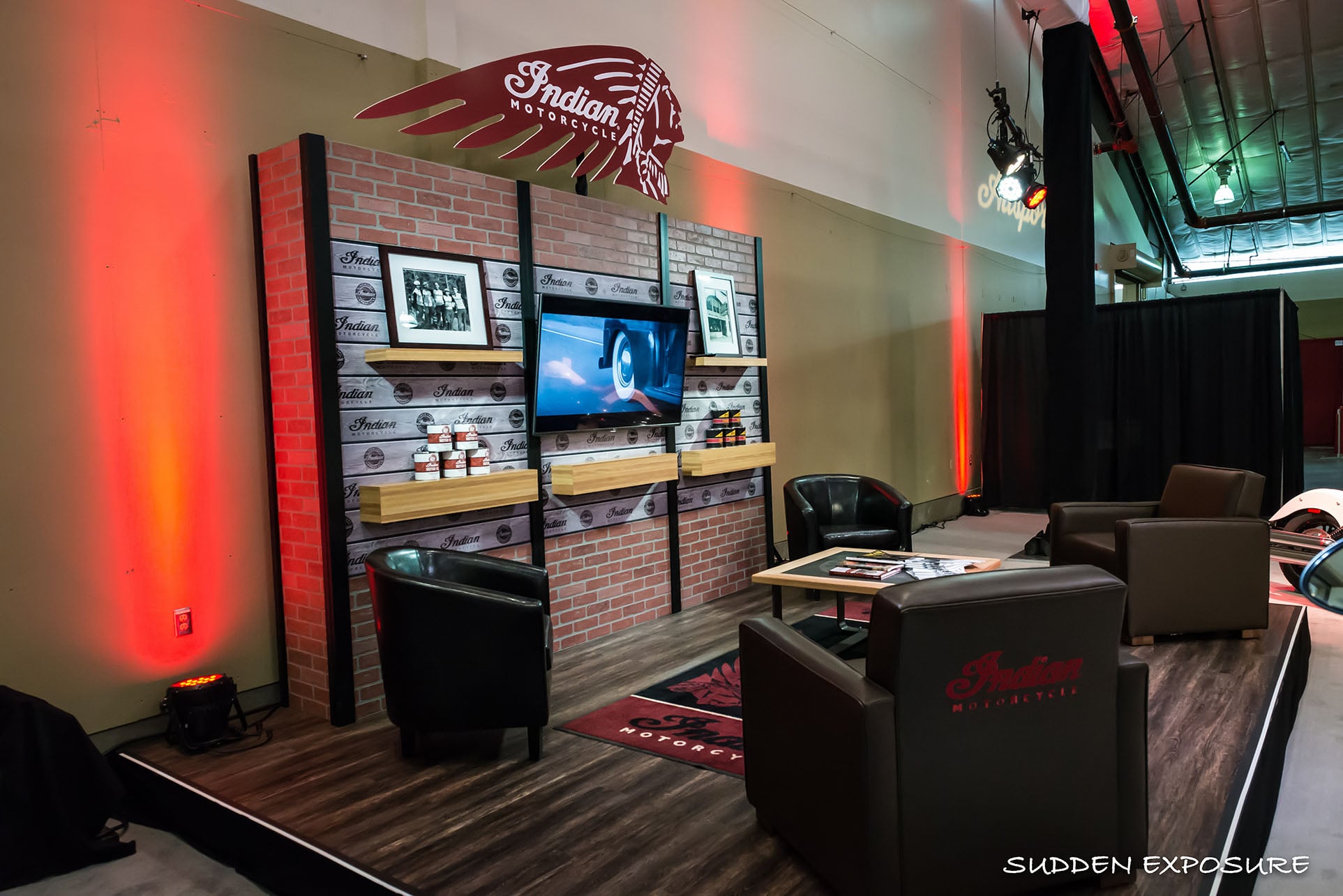 A branded motorcycle lounge with a brick-style display wall, screen, shelving and leather seating.