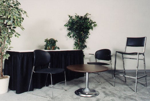 Black chairs and a barstool with a low round pedestal table, a skirted rectangular table and potted plants.