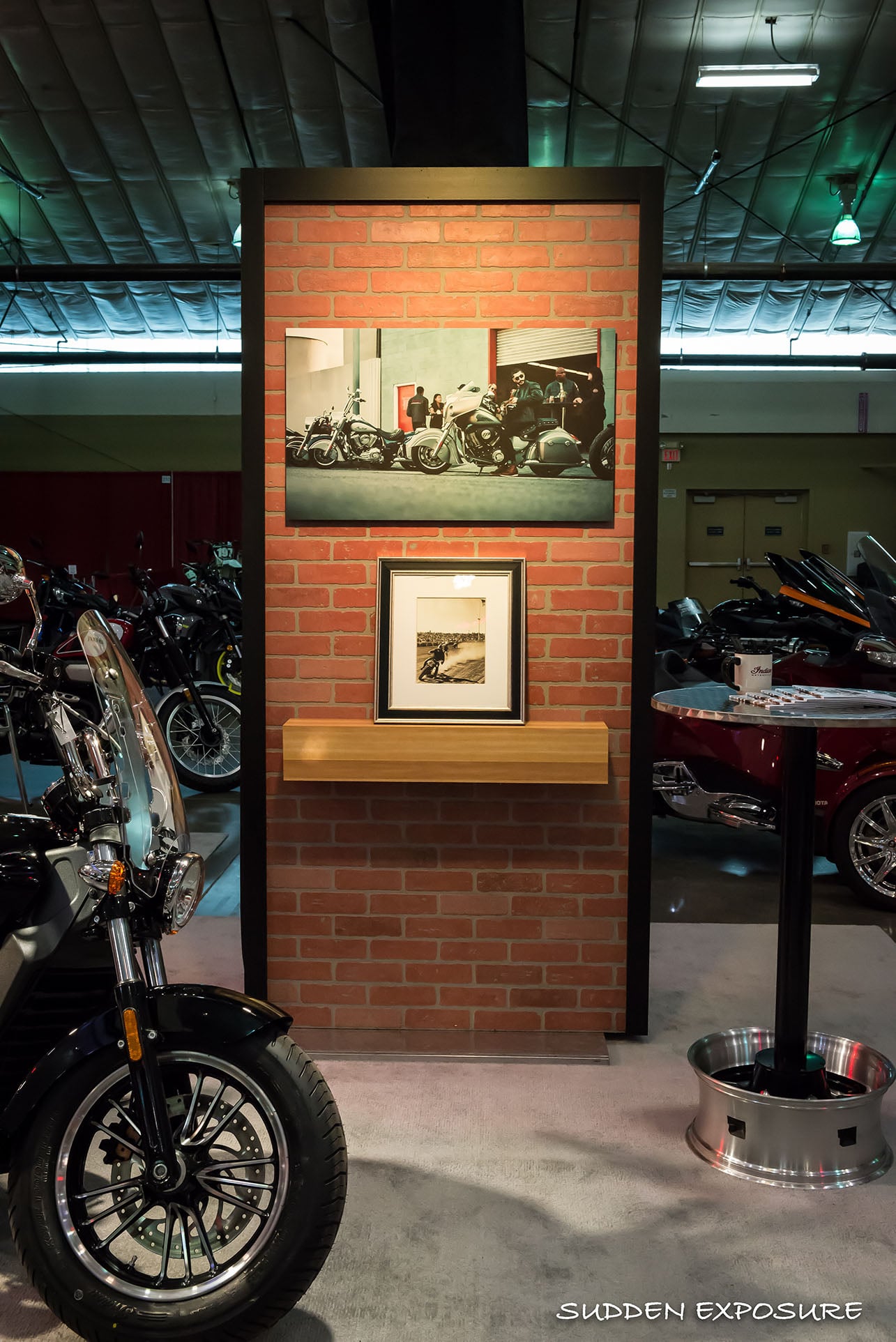 A brick-style display panel with framed motorcycle photography and a wood shelf.