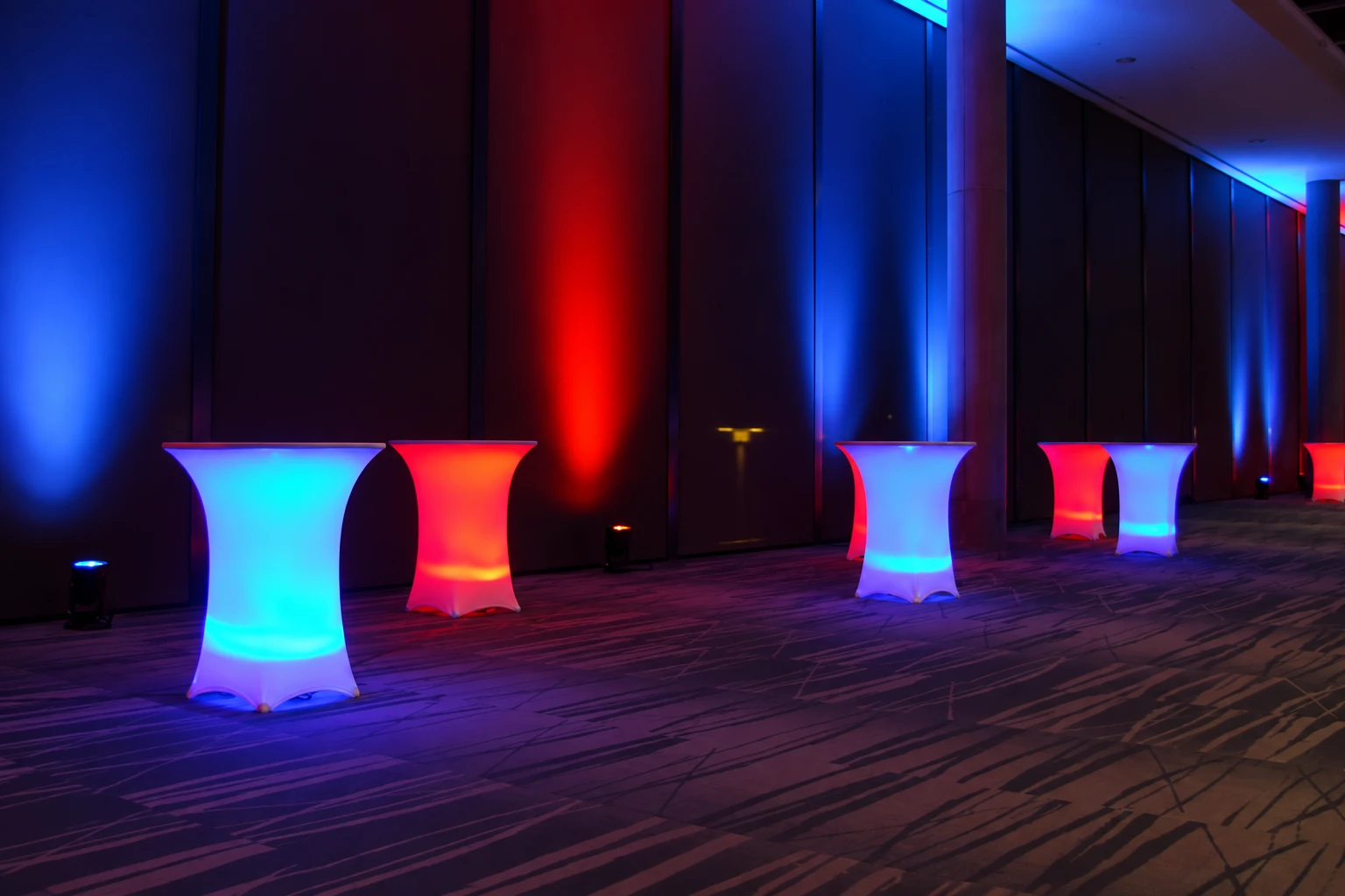 Blue and red illuminated spandex cocktail tables arranged along a wall with matching event uplighting.