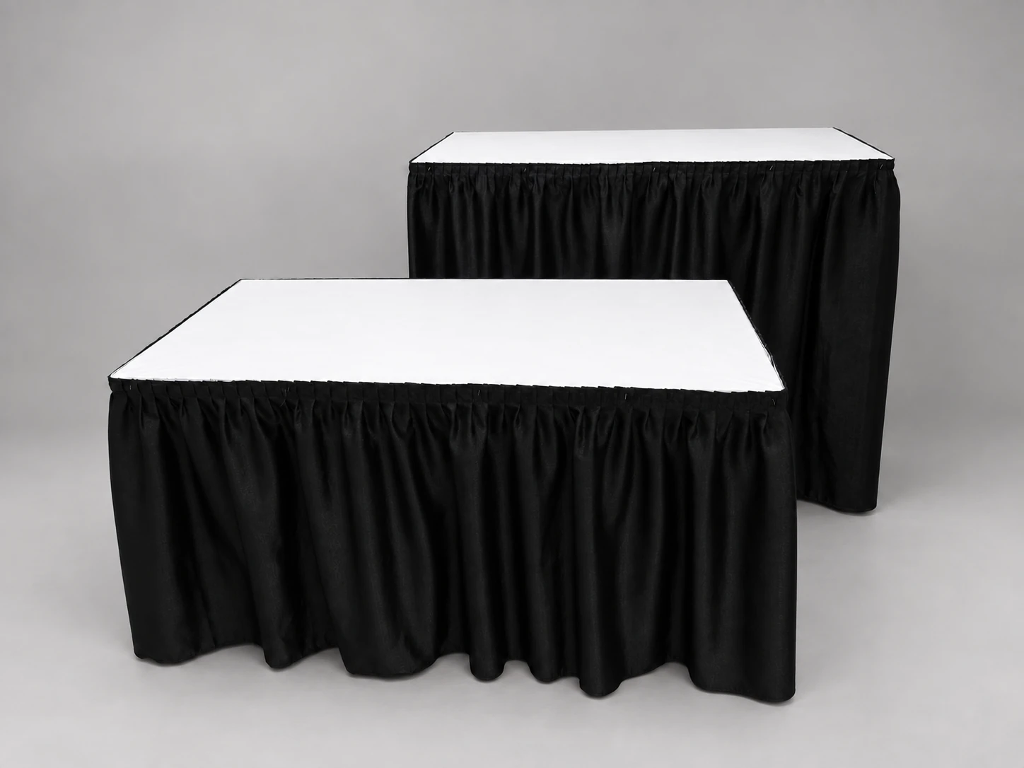 A standard-height rectangular table in front of a taller counter-height table, both with white tops and black skirts.